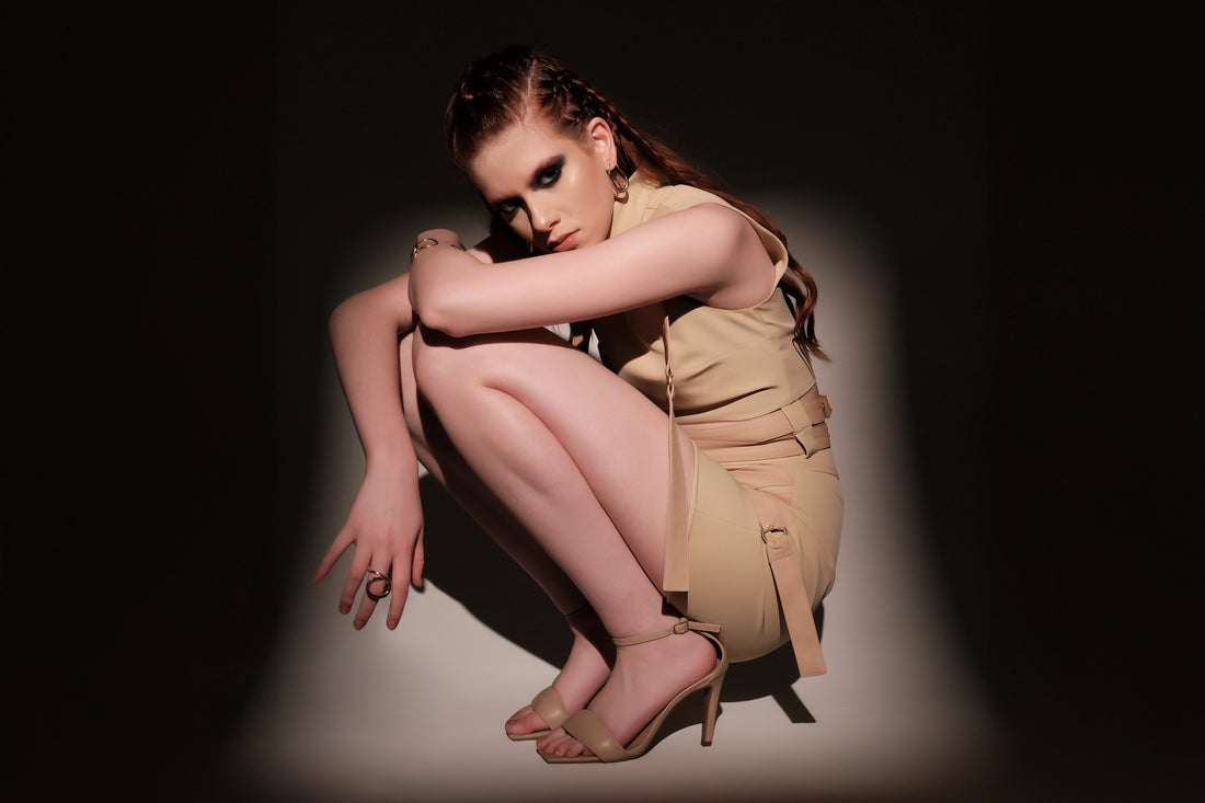 Model crouched in a beige harness skirt set, surrounded by spotlight shadows, sculptural and futuristic black and white fashion portrait.